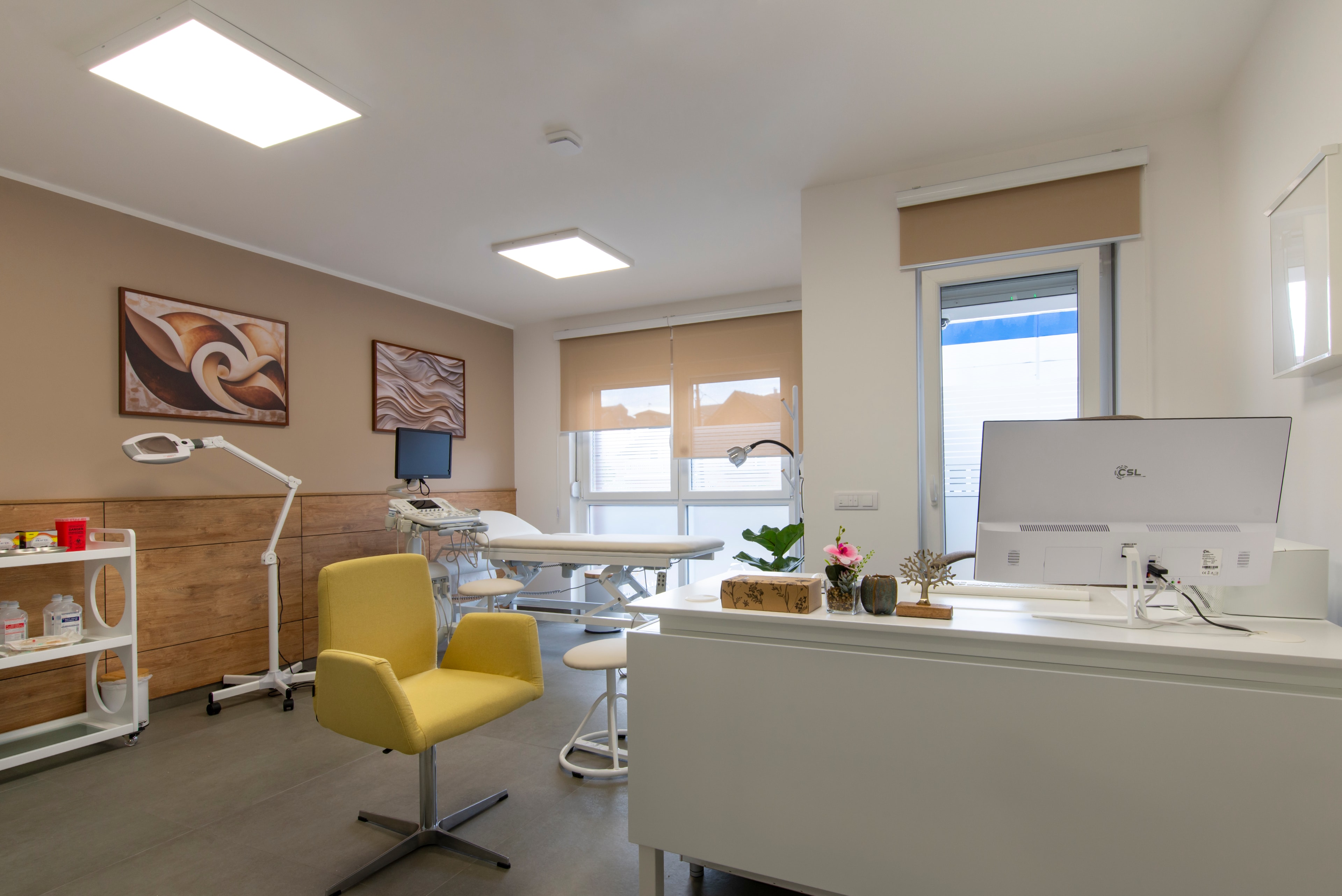 A bright, modern aesthetic clinic treatment room with a reclining treatment couch, magnifying lamp and consultation desk.